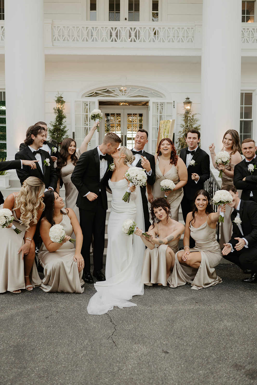 Bride and groom kiss while their wedding party reacts with playful faces in front of the mansion.